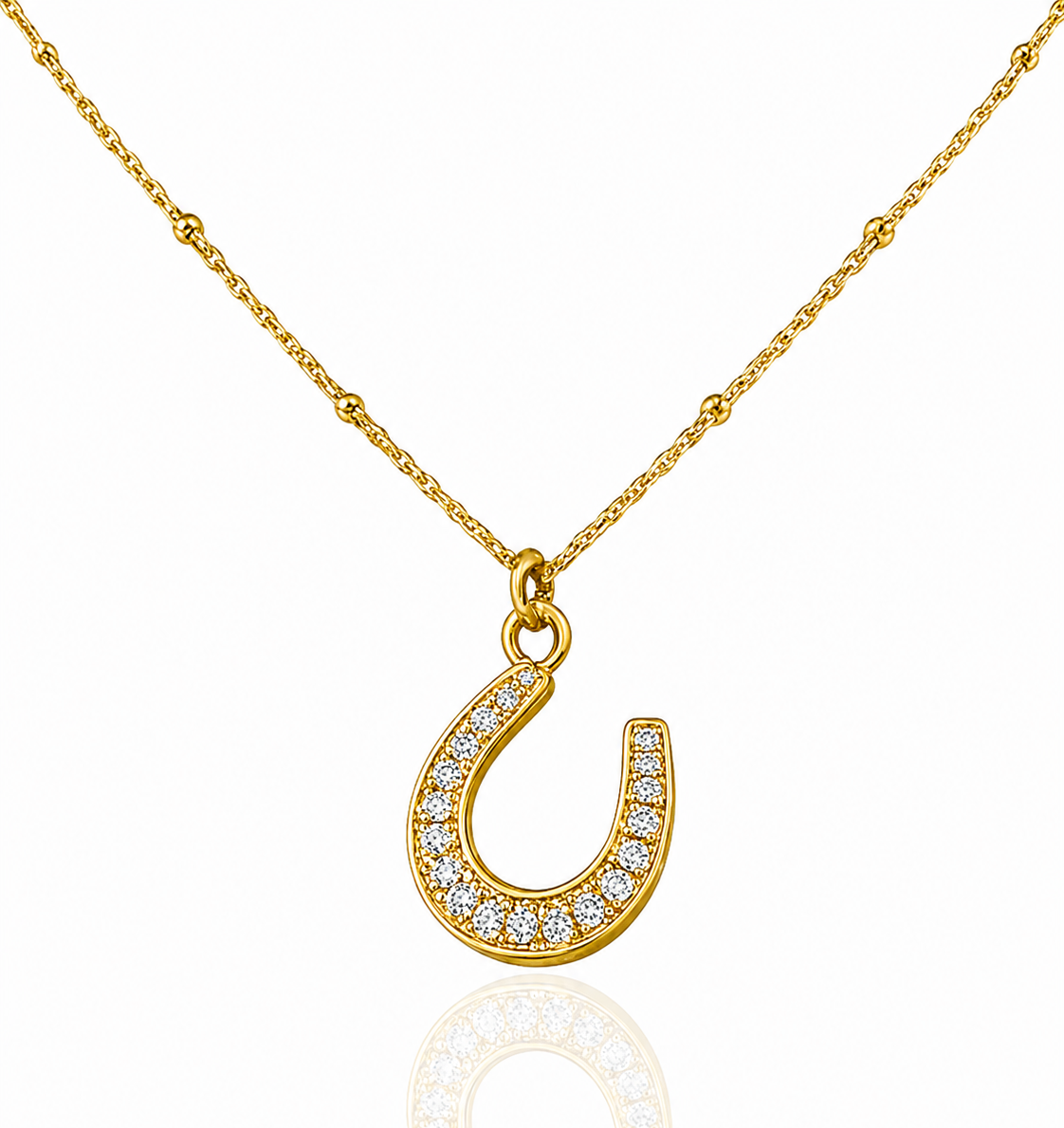 A gold horseshoe pendant necklace paved with small clear stones, suspended from a delicate gold chain on a white background with a soft reflection below.
