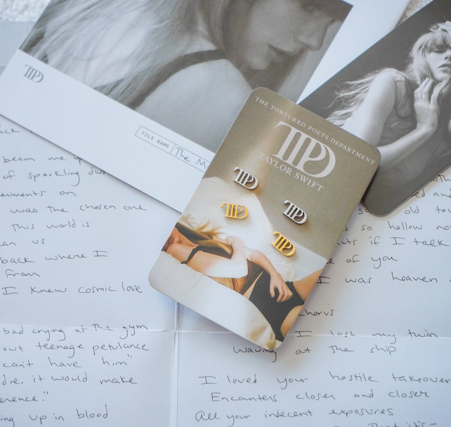 Taylor Swift 'The Tortured Poets Department' gold and white stud earrings on their original display card, styled on top of handwritten letters.