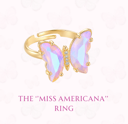 A gold butterfly ring with iridescent pastel purple-blue-green opal-like wings and a small clear stone in the center, shown on a pale pink background with the text 'THE "MISS AMERICANA" RING' and 'www.hebejewel.