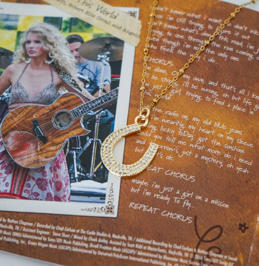 A gold horseshoe pendant necklace paved with small clear stones, displayed on a brown printed collage background featuring song lyrics and a photo of a woman playing guitar.