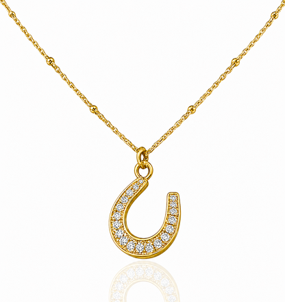 A gold horseshoe pendant necklace paved with small clear stones, suspended from a delicate gold chain on a white background with a soft reflection below.