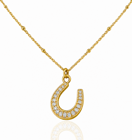 A gold horseshoe pendant necklace paved with small clear stones, suspended from a delicate gold chain on a white background with a soft reflection below.