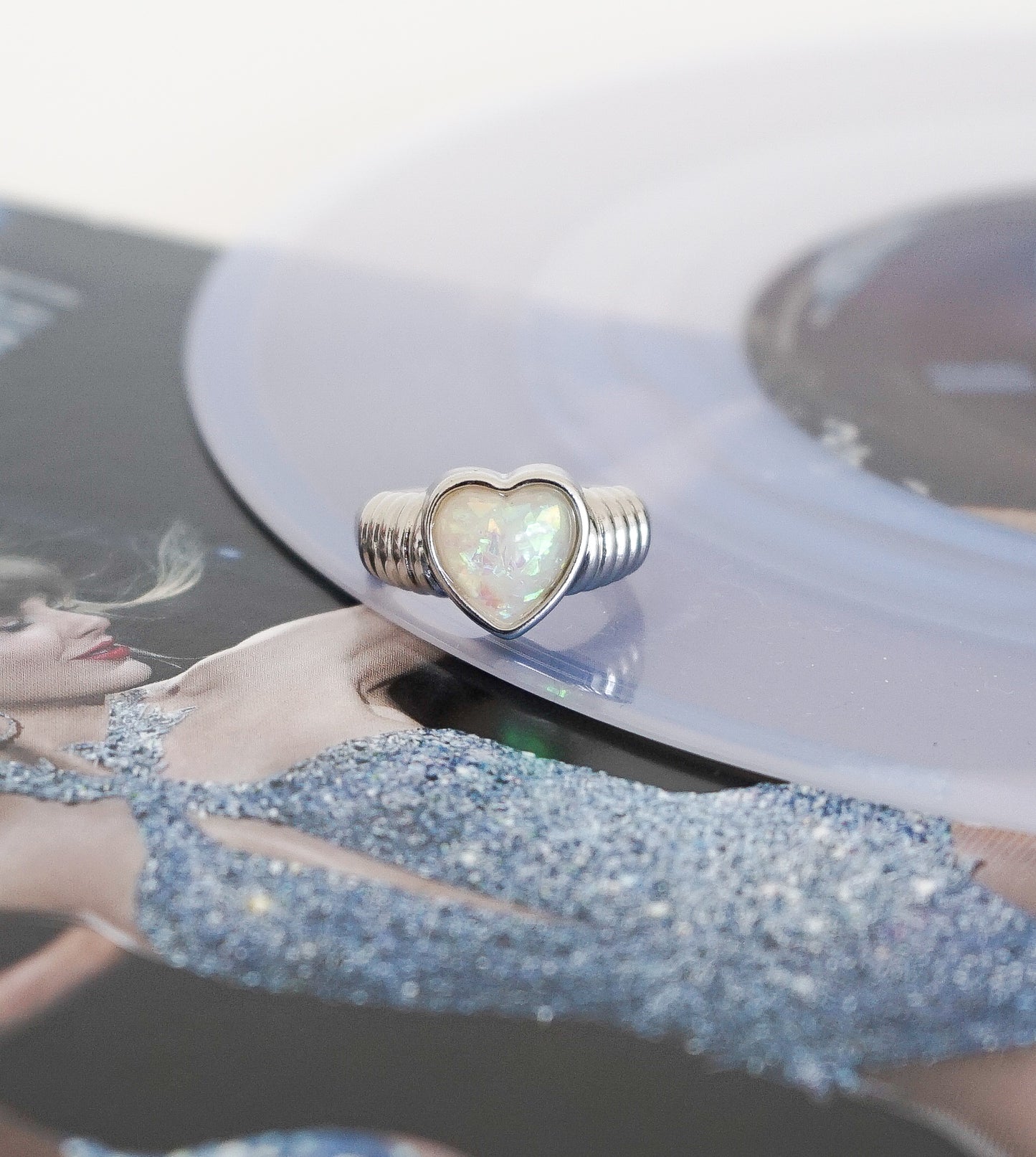 A silver ring with a heart-shaped iridescent opal center, resting on a translucent vinyl record over a printed photo of a woman in a blue glitter dress.