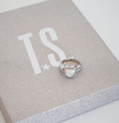 A silver ring with a heart-shaped iridescent opal inlay, resting on a glittery silver presentation box with white 'TS' lettering.