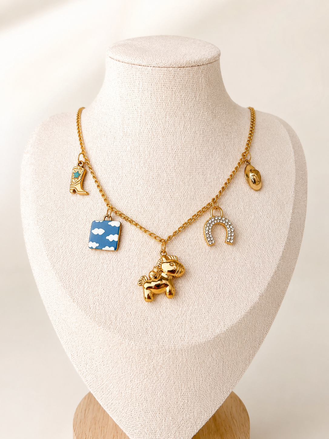 A gold-tone charm necklace displayed on a cream fabric jewelry bust. The necklace features a central gold unicorn charm, a blue square charm with white clouds, a gold horseshoe with clear rhinestones, and two small gold animal charms.